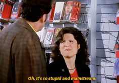 a woman talking to a man in front of a book shelf with books on it