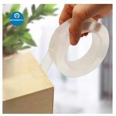 a hand holding a clear tape over a wooden block with a plant in the background