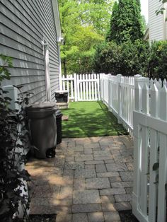 a white picket fence in front of a house with a black trash can on the sidewalk