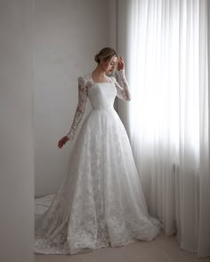 a woman in a white wedding dress standing next to a window with sheer drapes