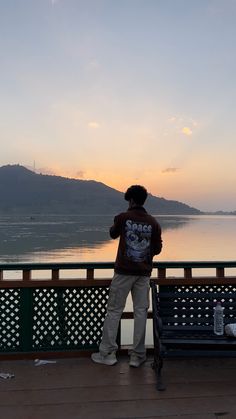 a man standing on top of a wooden deck next to a lake at sunset with mountains in the background