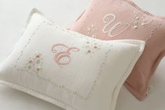 two pillows with embroidered letters on them sitting next to each other, one pink and the other white