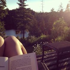 a person reading a book while sitting on a bench in front of a lake and forest