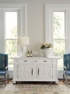 two blue chairs and a white cabinet with wine bottles on it in front of windows