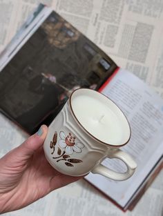 a hand holding a coffee cup with a candle in it next to an open book