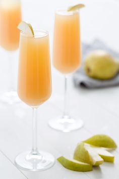 three glasses filled with orange juice and limes on a white tablecloth next to some fruit