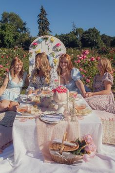 four women sitting at a table with food and drinks in front of them, outdoors