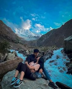 a man and woman sitting on top of a rock next to a mountain river while holding each other