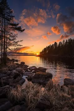 the sun is setting over a lake with rocks and grass in front of it,