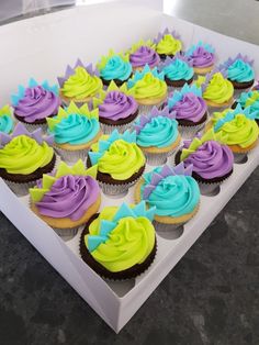 there are many cupcakes in the box on the table and one is decorated with blue, purple, and green icing