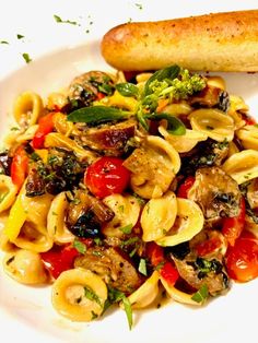 a white plate topped with pasta and veggies next to a baguette