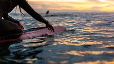 a woman kneeling on a surfboard in the ocean at sunset or dawn, with her hand over the edge of the board