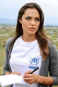 a beautiful young woman holding a pen and paper in her hand while standing on top of a hill