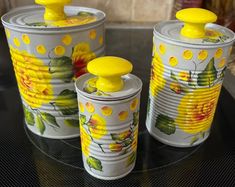 three tin canisters with yellow flowers painted on them sitting on a glass table