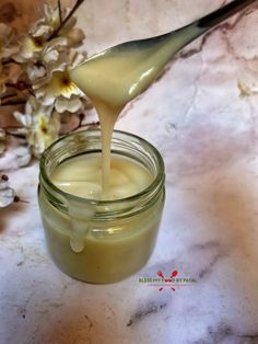 a spoon pouring honey into a jar on a marble surface with flowers in the background