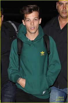 the man is wearing a green hoodie and black pants with his hands in his pockets