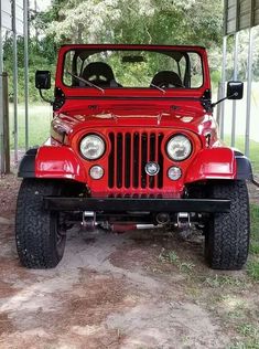 a red jeep parked in front of a metal structure with trees and grass behind it