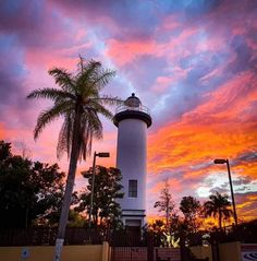 a white light house sitting next to a palm tree under a colorful sky at sunset