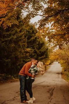 a man and woman kissing in the middle of an empty road surrounded by autumn trees