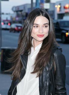 a woman with long brown hair wearing a black leather jacket and white shirt is walking down the street