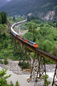 a train traveling over a bridge next to a forest filled mountain side on top of a river