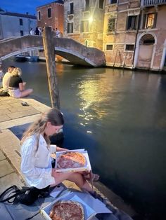 a woman sitting on the edge of a canal with two pizzas in front of her