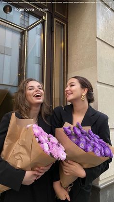 two women are holding flowers and laughing outside the door of a building with their hands in each other's pockets