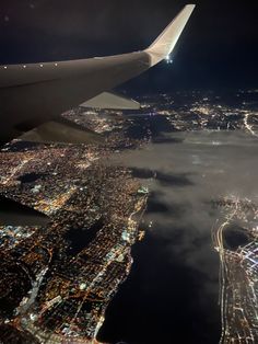 the wing of an airplane flying over a city at night with lights and clouds in the foreground