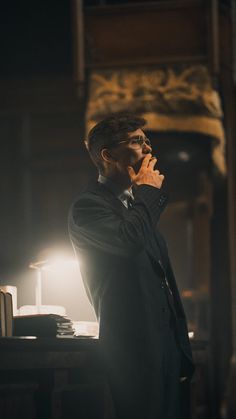 a man in a suit standing next to a table with a lamp on it and his hand near his mouth