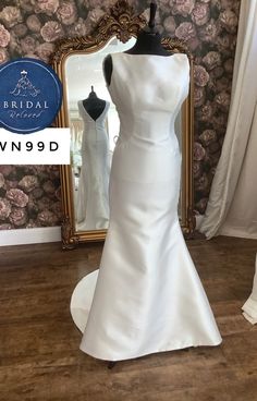 a white wedding dress on display in front of a floral wallpapered room with a gold framed mirror