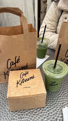 two green drinks are sitting on the table next to brown paper bags with black writing