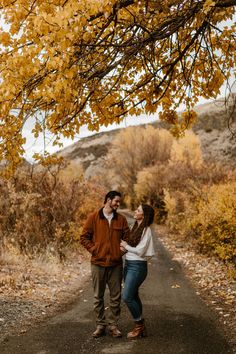 a man and woman walking down a dirt road under a tree with yellow leaves on it