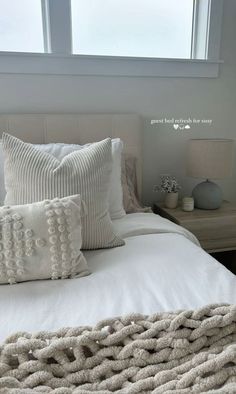 a bed with white pillows and blankets on it next to a night stand in front of a window