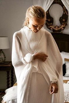 a woman wearing a white dress standing in front of a bed