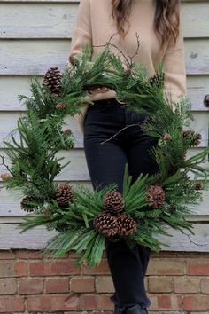 a woman standing in front of a building holding a wreath with pine cones on it