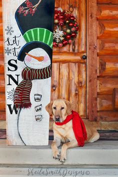 a dog sitting next to a snowman painted on a wooden sign