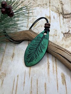 a green leaf shaped pendant hangs from a leather cord on a wooden table next to pine cones