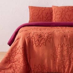 an orange comforter and two pillows on a bed with wood flooring in front of a white wall