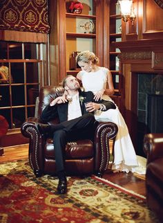 a bride and groom sitting on a leather chair in front of a fireplace at their wedding