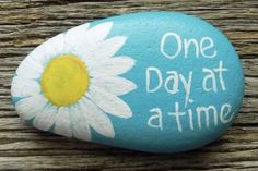 a painted rock that says one day at a time with a white daisy on it