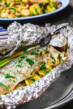 fish and vegetables wrapped in tin foil on a plate with other food dishes behind it