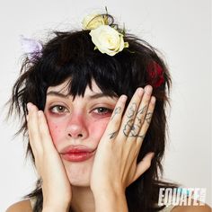 a woman with tattoos holding her hands to her face and looking at the camera while wearing a flower in her hair