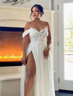 a woman standing in front of a fireplace wearing a white dress with thigh high slit