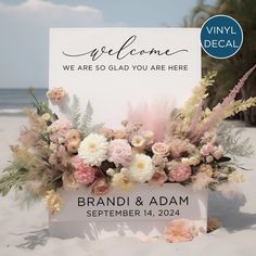 a welcome sign on the beach with pink and white flowers in front of it that reads, we are so glad you are here