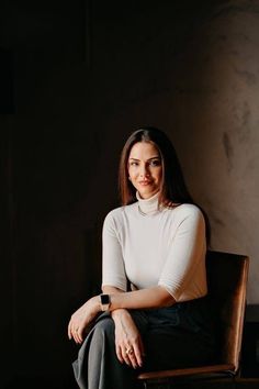 a woman sitting in a chair with her arms crossed and looking at the camera while wearing a white shirt