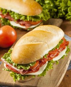 two sub sandwiches sitting on top of a wooden cutting board next to tomatoes and lettuce