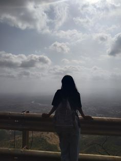 a woman standing on top of a wooden fence next to a lush green hillside under a cloudy sky