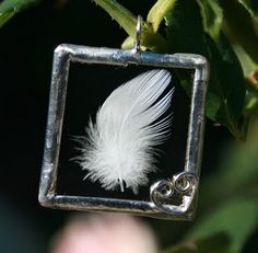 a white feather is hanging from a silver square pendant on a tree branch with leaves in the background