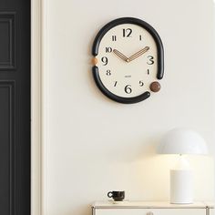 a clock mounted to the side of a white wall next to a table with a lamp on it