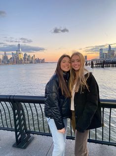 two young women standing next to each other near the water in front of a city skyline